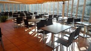Un amplio espacio exterior para comer con mesas y sillas, ventanas grandes y suelo de baldosas.
