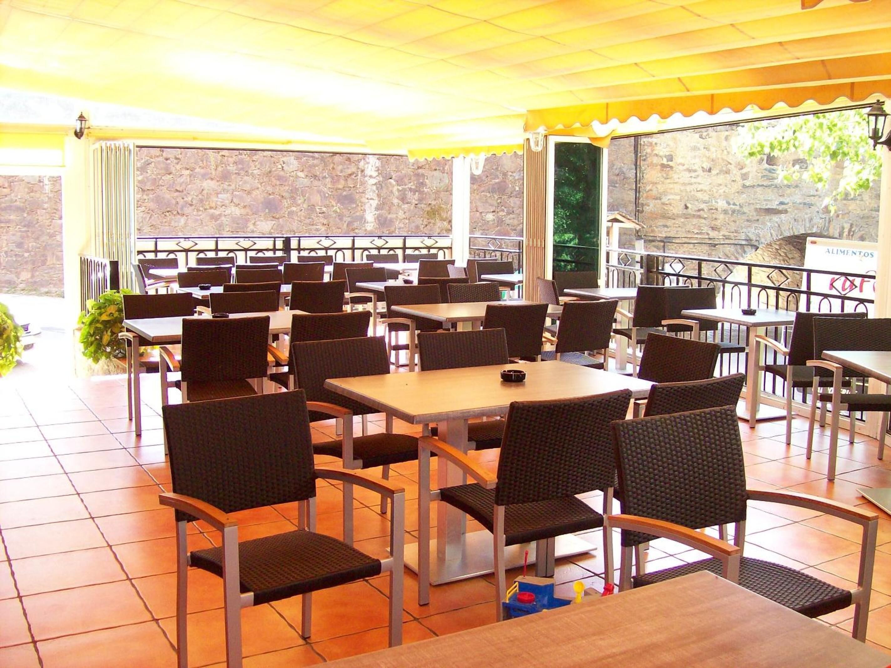 Un espacio de comedor al aire libre cubierto con varias mesas y sillas, un muro de piedra al fondo y un letrero que dice 'ALIMENTOS'.
