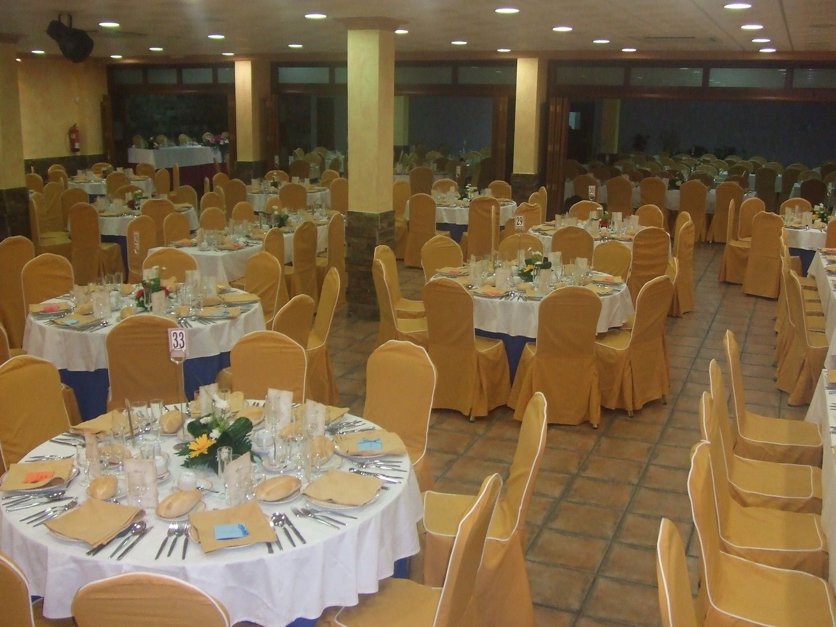 Un salón de banquetes con mesas redondas, cada una cubierta con manteles blancos, sillas doradas y una vajilla elegantemente dispuesta.