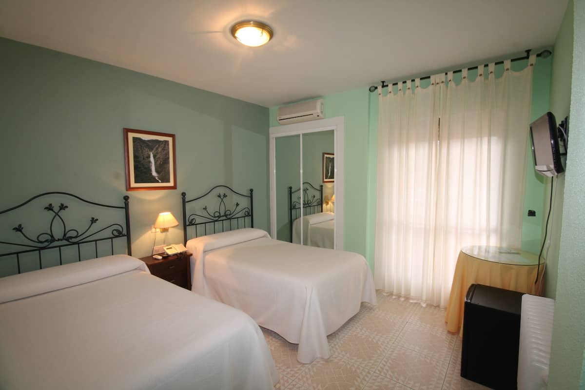 Una habitación de hotel con dos camas, una mesita de noche con lámpara, televisión y una ventana con cortinas.