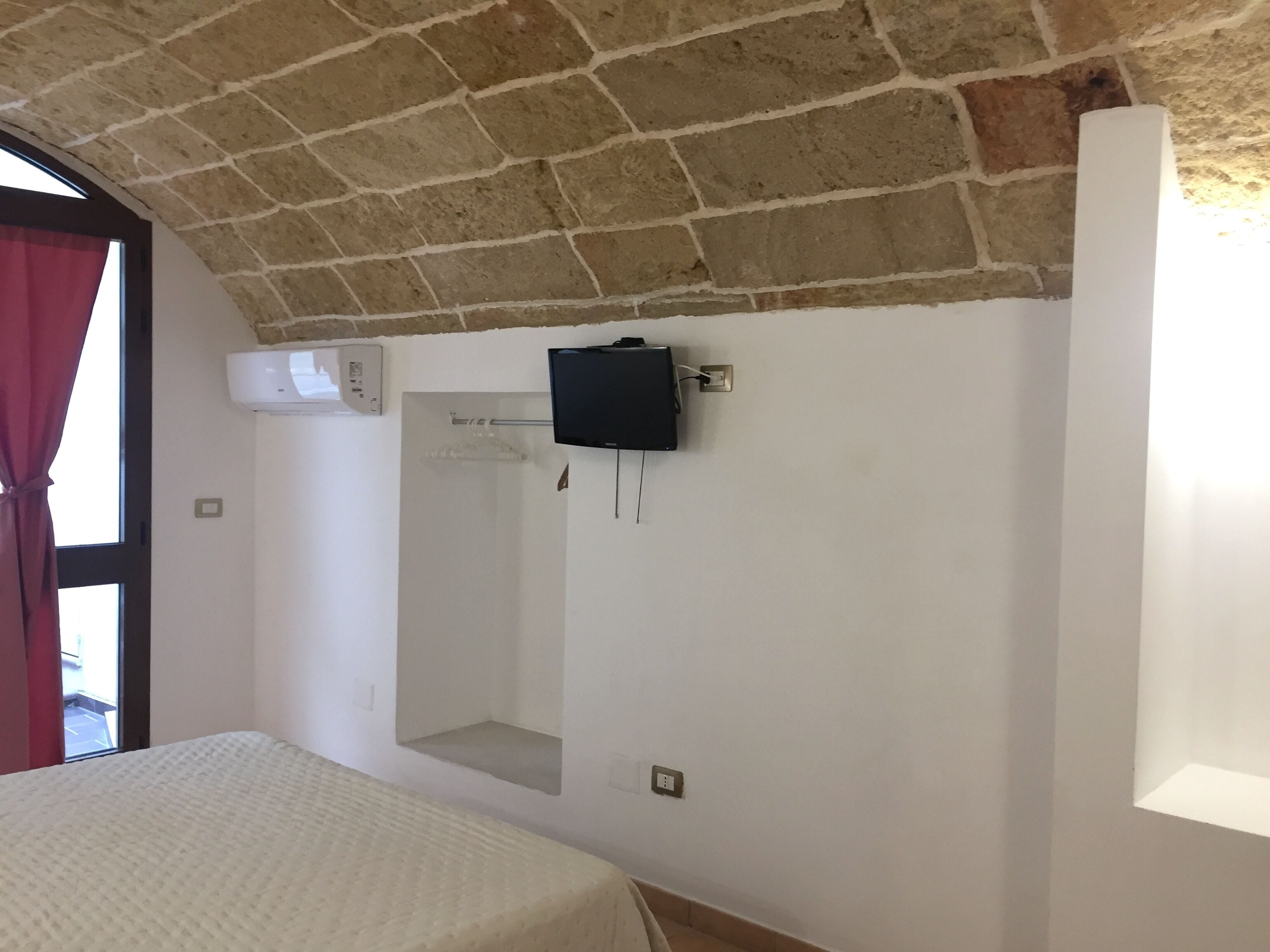 A bedroom with a brick ceiling, a mounted TV, a window with red curtains, and a bed with a white cover.