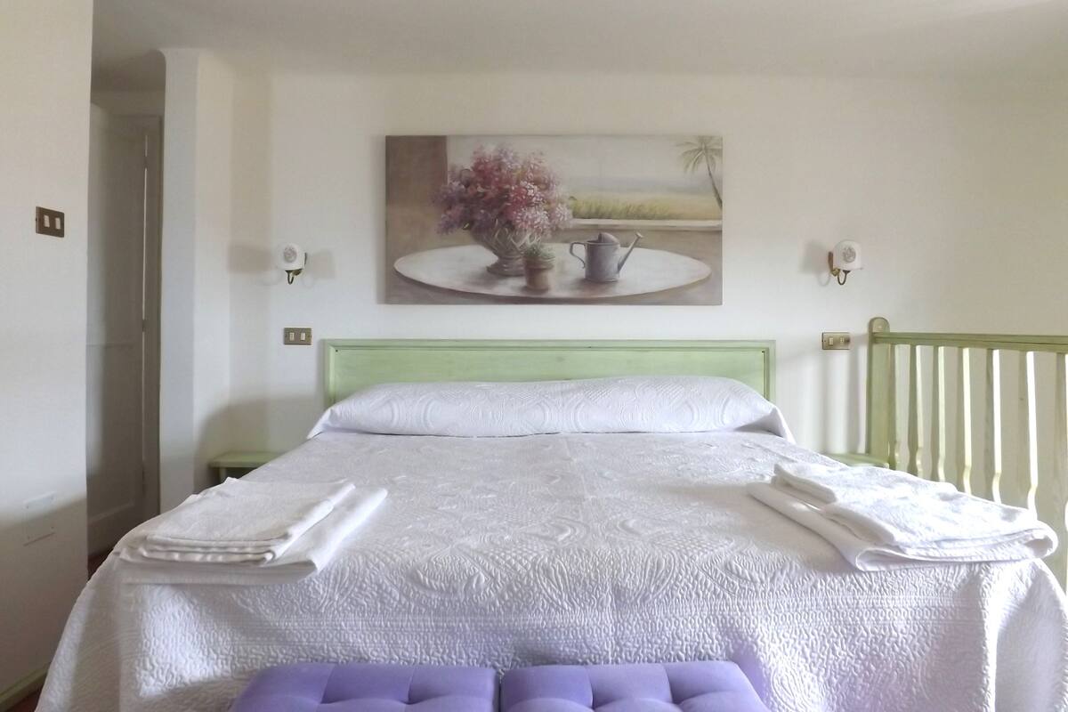 A neatly made bed with a white duvet, a purple cushion, and a folded towel on top. There is a painting of flowers and a teapot on the wall above the bed.