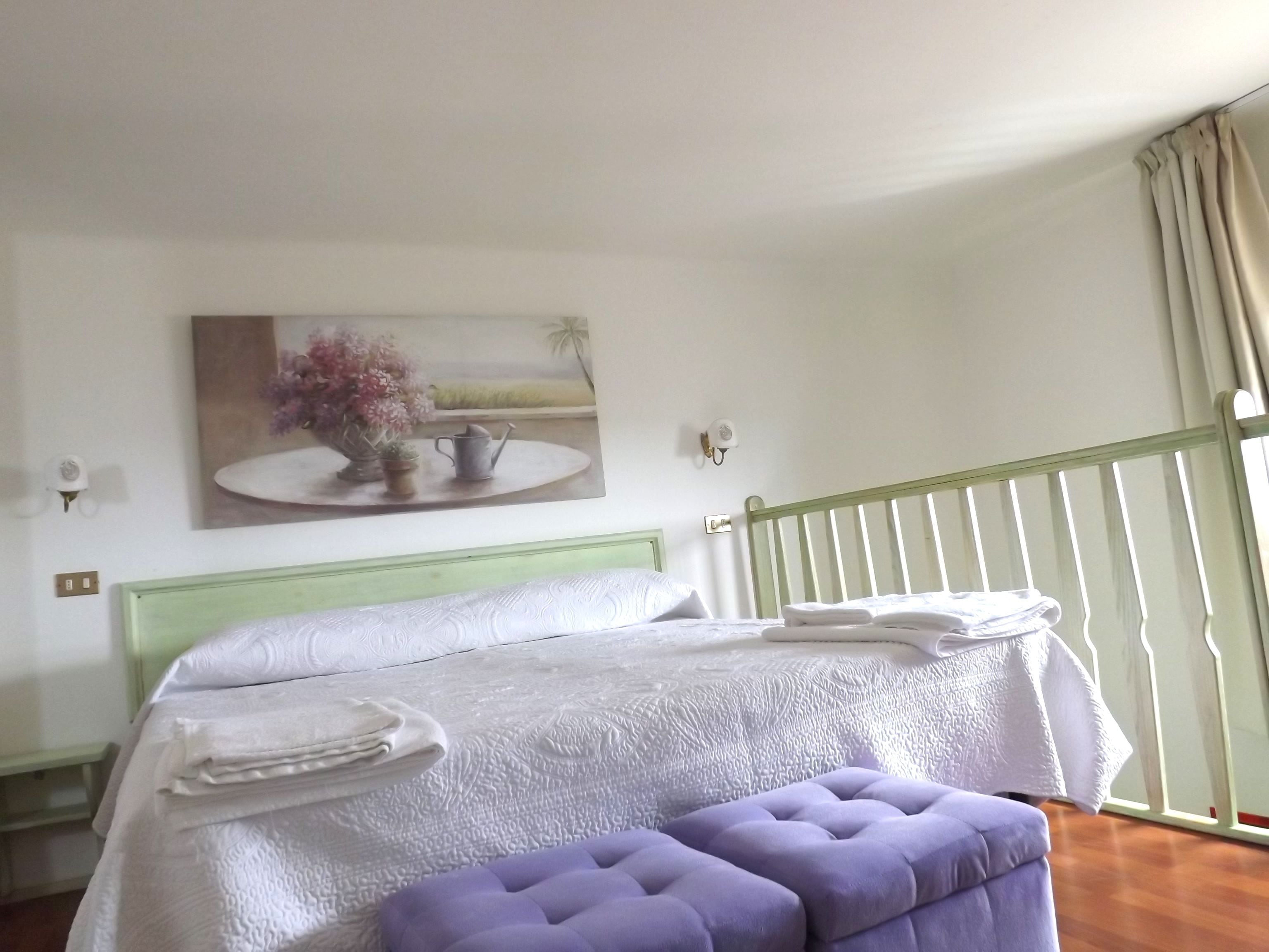 A hotel room with a bed, a green headboard, a painting on the wall, and a balcony with curtains.