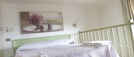 A hotel room with a bed, a green headboard, a painting on the wall, and a balcony with curtains.