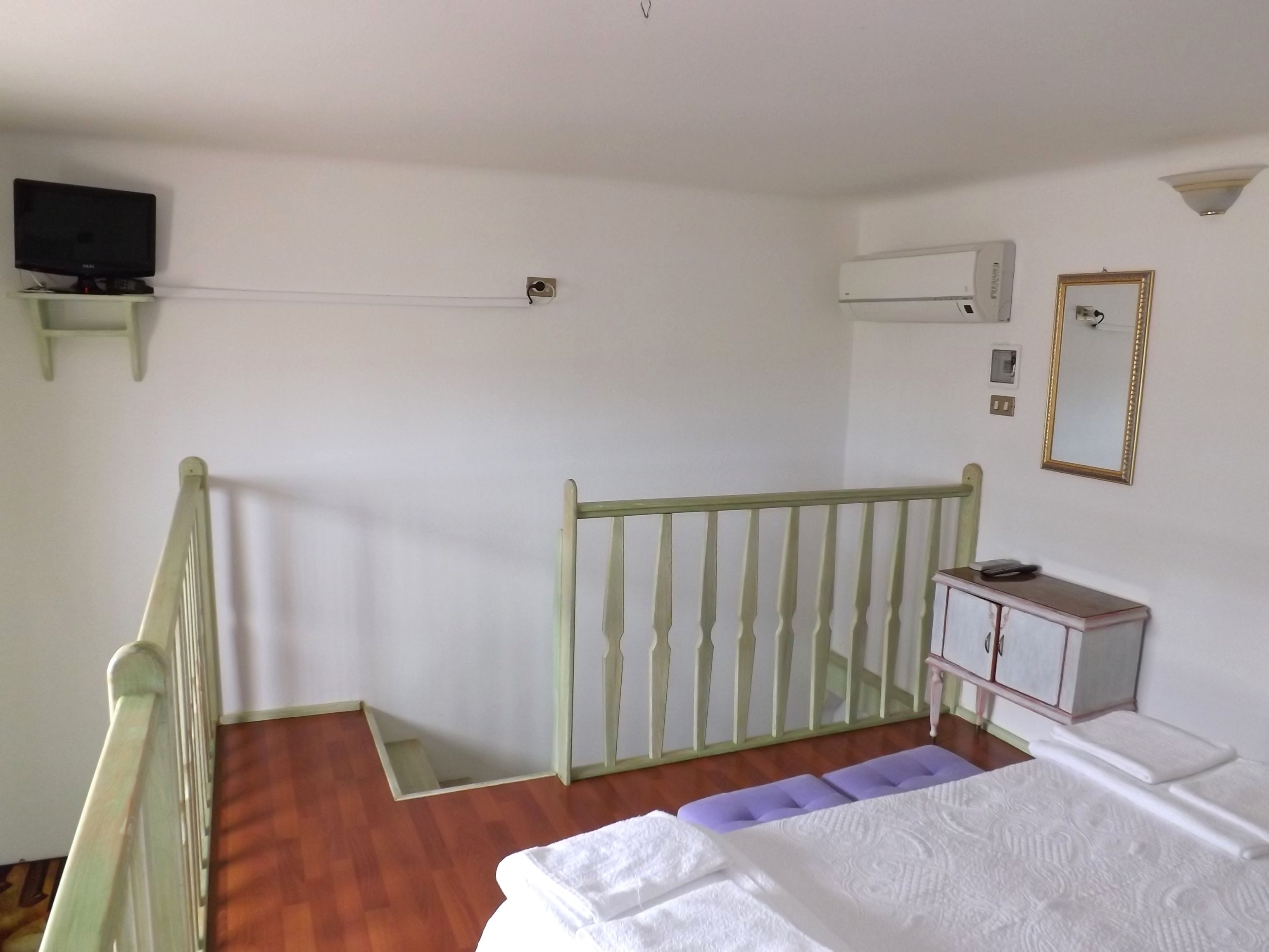 Ein Zimmer mit Holzboden, einem Bett mit weißer Bettwäsche, einem kleinen Tisch mit Spiegel und einem Fernseher auf einem Regal.