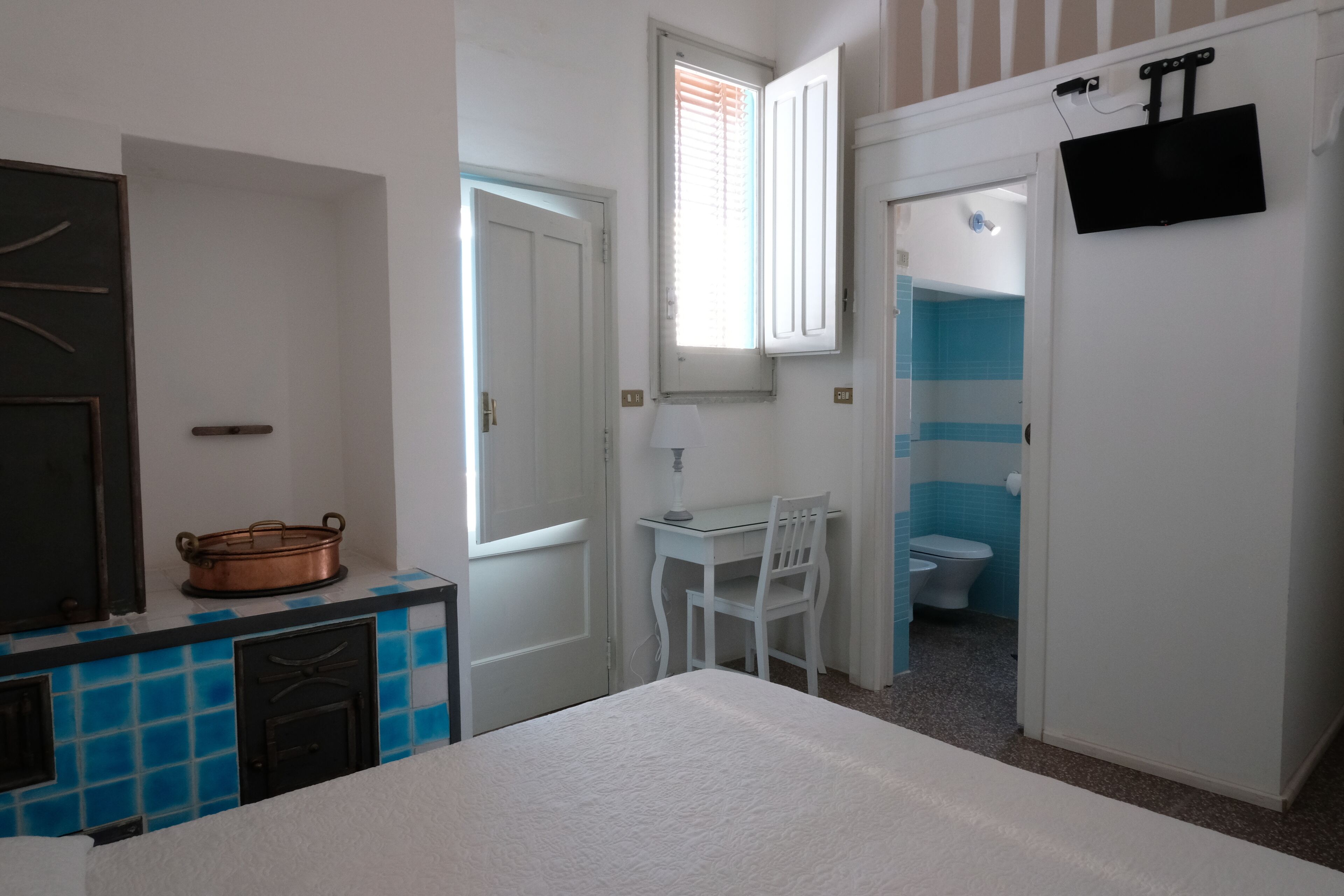 A room with a bed, a desk with a chair, a window with blinds, a white toilet, and a TV mounted on the wall.