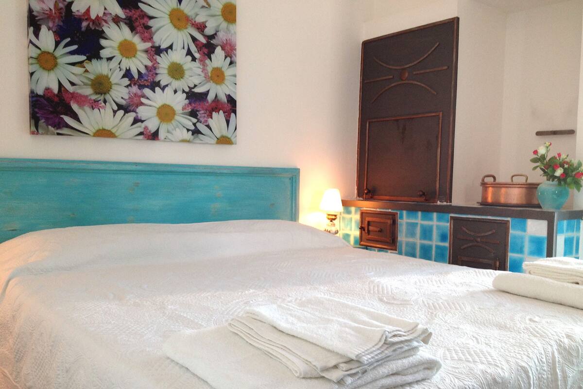 A neatly made bed with folded towels, a blue headboard, a wall-mounted painting of daisies, and a small table with a vase of flowers.