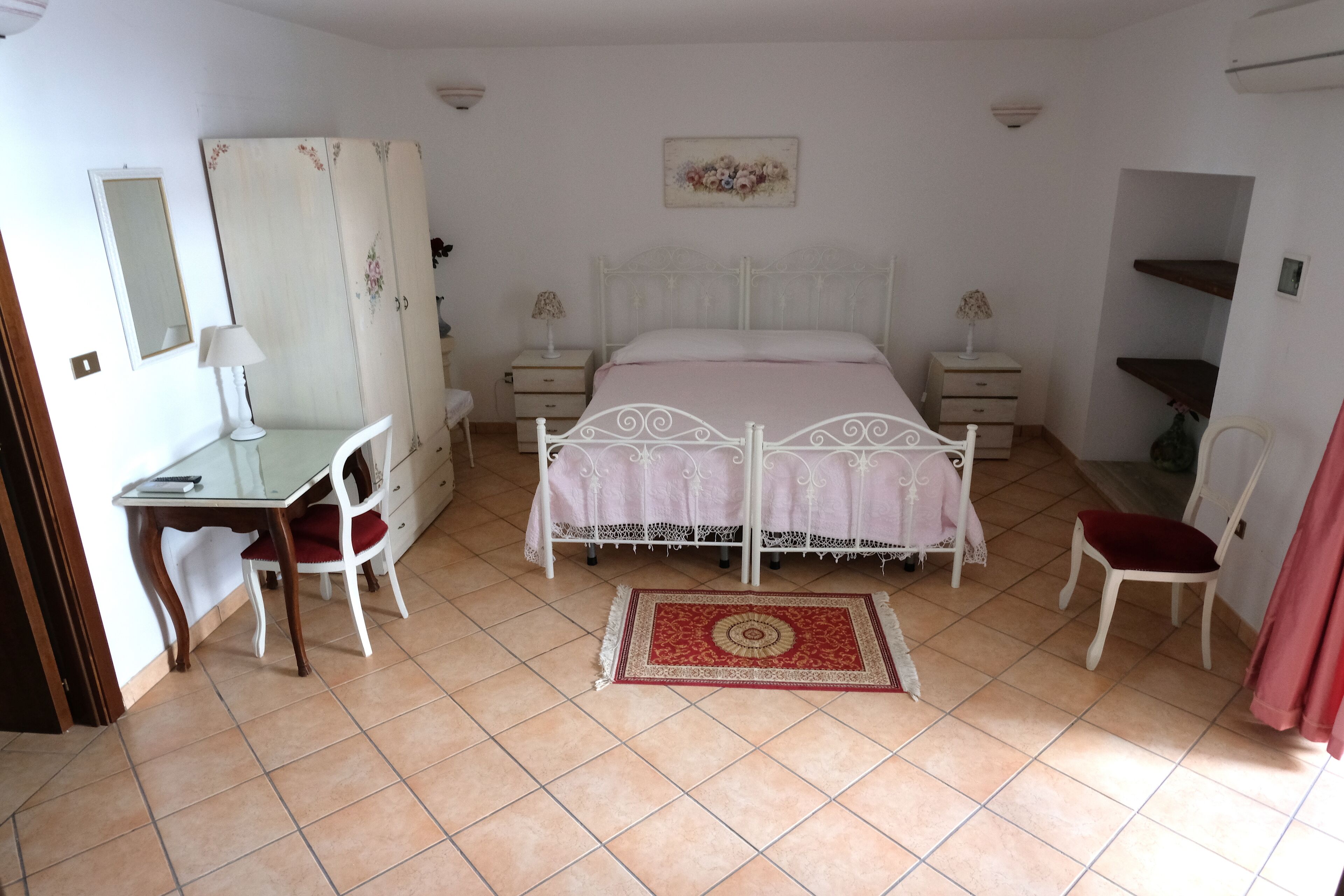 A bedroom with a bed, a desk, a chair, a wardrobe, and a small table with a lamp.