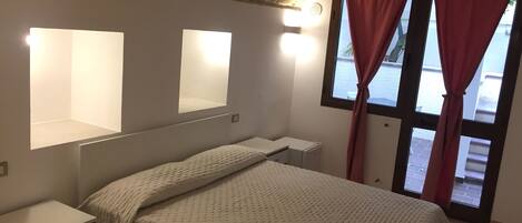 Quarto com cama, mesas de cabeceira, espelho e janela com cortinas.