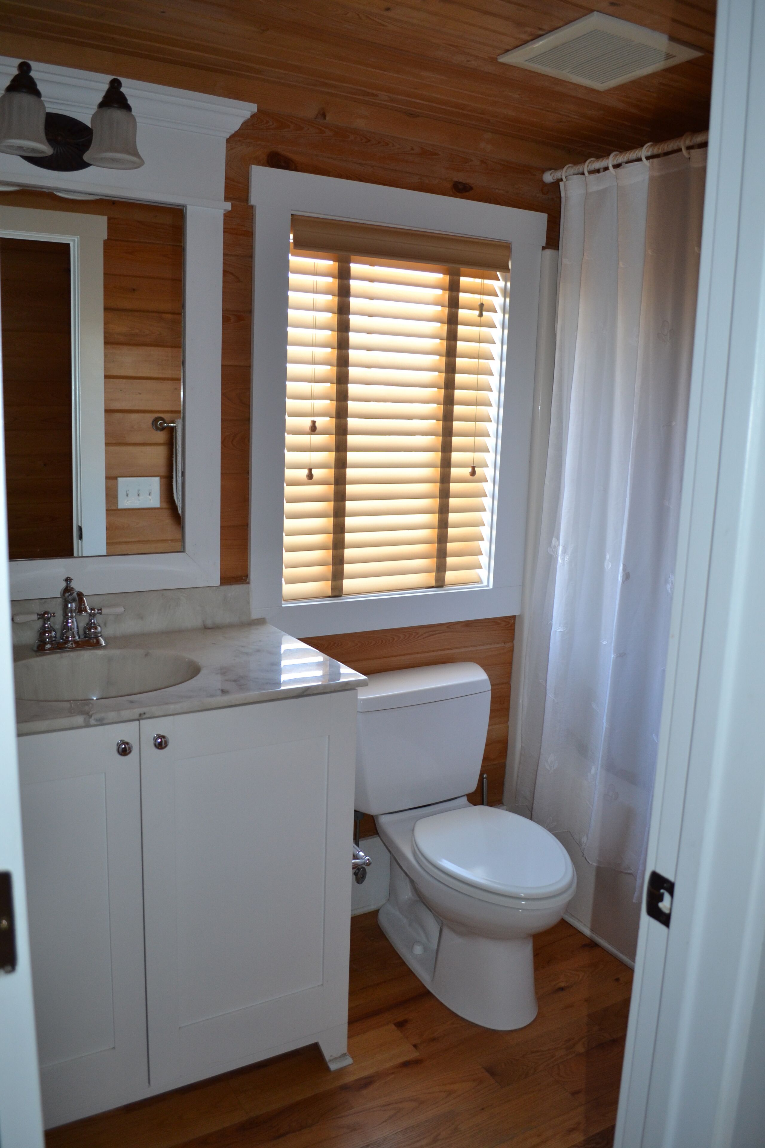 Une salle de bain avec un plancher en bois, des accessoires blancs, une fenêtre avec des stores et un rideau de douche.