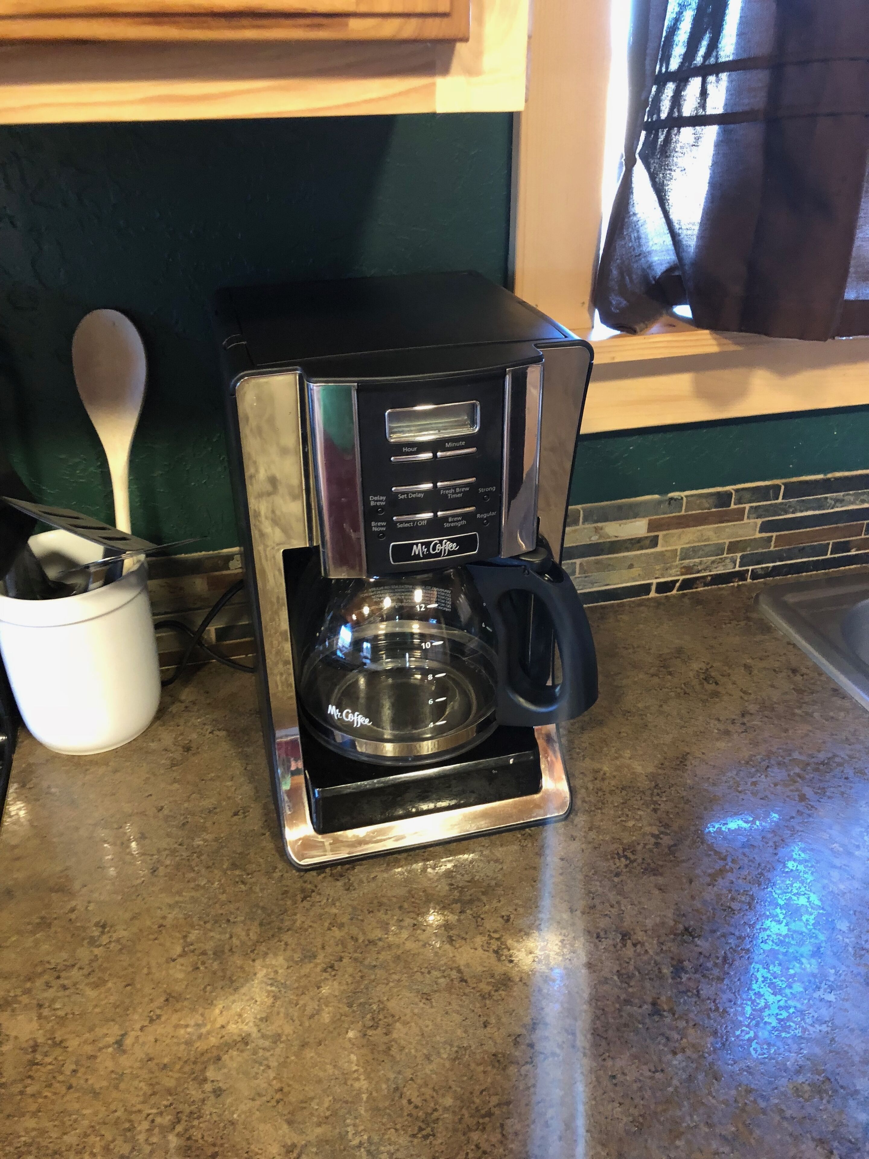 A coffee maker on a kitchen counter with a spoon and a white mug nearby.