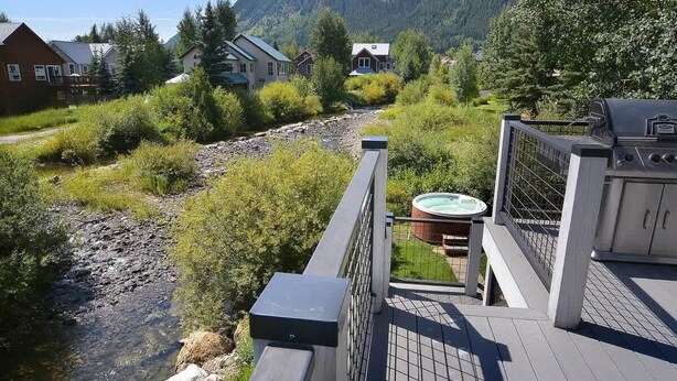 A deck with a hot tub, a grill, and a view of a river and houses in a forested area.