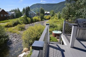 A deck with a hot tub, a grill, and a view of a river and houses in a forested area.