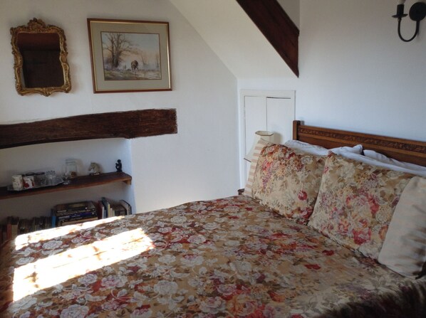 A bedroom with a floral bedspread, a wooden headboard, a shelf with various items, and a framed picture on the wall.