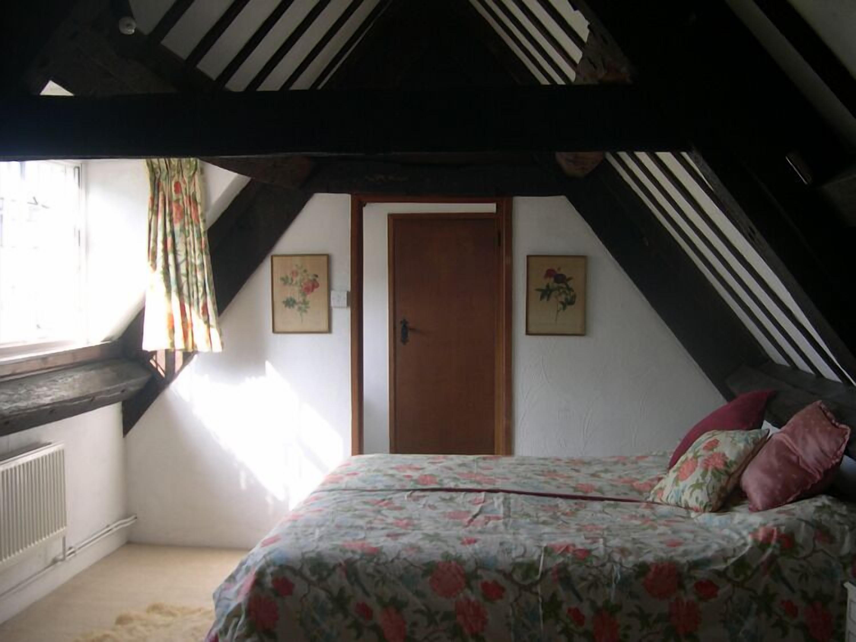 Un dormitorio con techo inclinado, una cama con ropa de cama floral, dos cuadros en la pared y una ventana con cortinas.