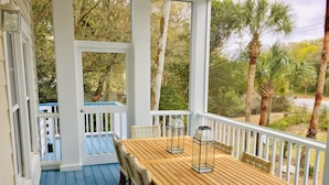 Una terraza de madera con una mesa y sillas, bordeada por barandas blancas, con vistas a un jardín con árboles y una piscina azul.