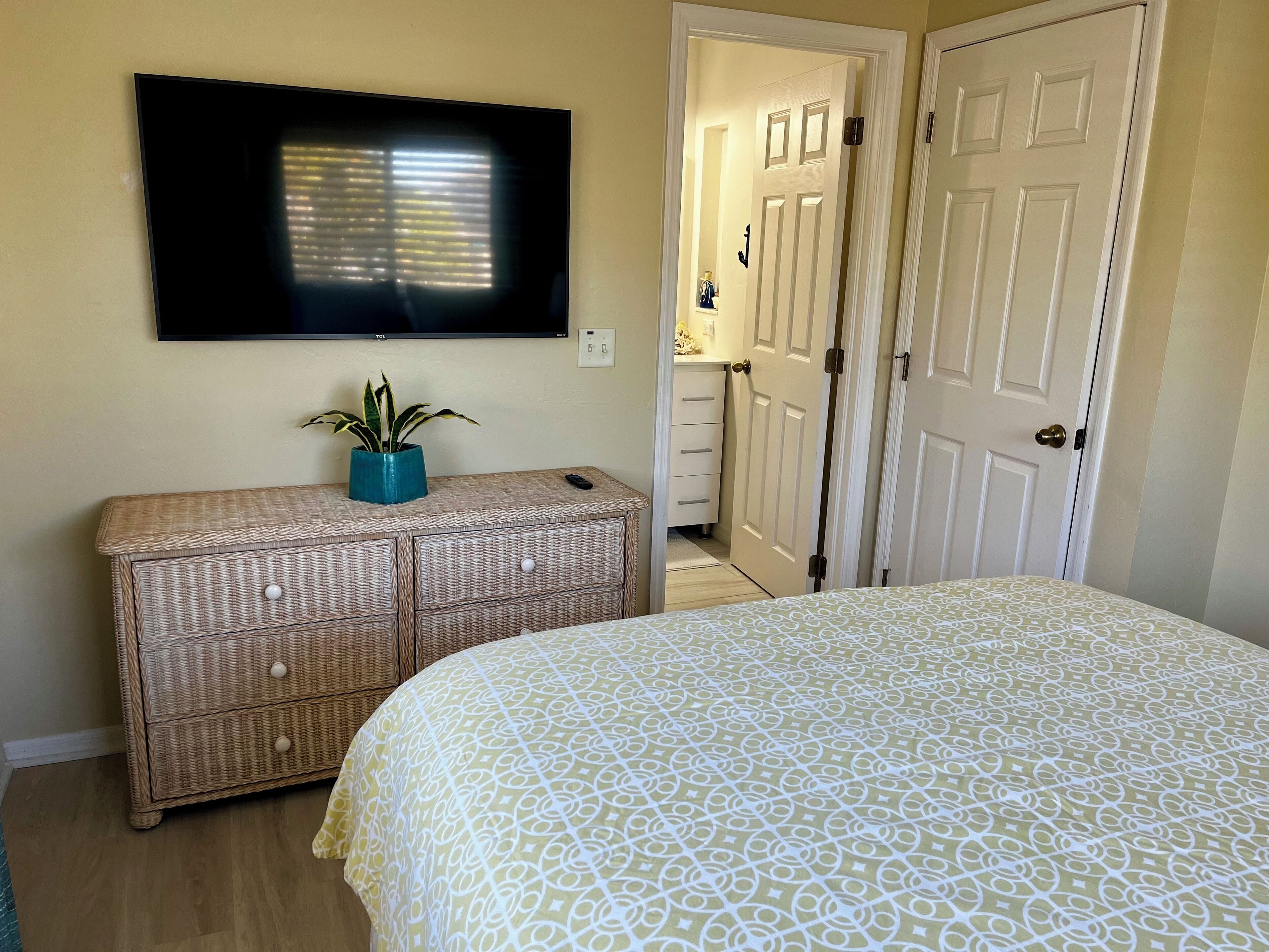 A bedroom with a bed, a television mounted on the wall, a dresser with drawers, and a door leading to another room.