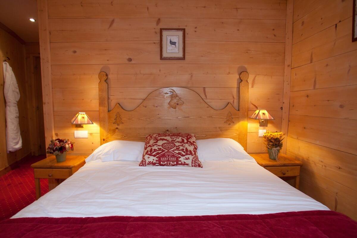 Une chambre à coucher aux murs lambrissés, avec un lit, deux lampes de chevet, un tableau encadré et un plaid rouge.