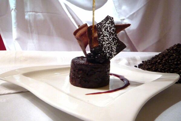 Un postre en un plato blanco con pastel de chocolate, un trozo de chocolate con confetis blancos y salsa roja.