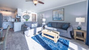 A modern living room with a grey sofa, a wooden coffee table, and a blue rug. There is a kitchen area with white cabinets and a ceiling fan. - The Summit 1BR 1.5BA Beautiful unit with great views! (Panama City Beach)