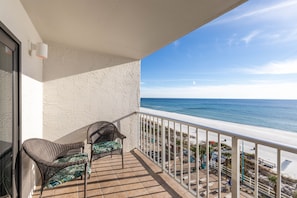 A balcony with wicker chairs and a table, overlooking a beach with a clear sky.
