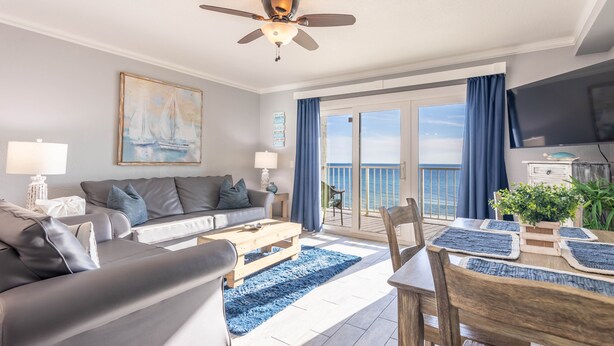 A modern living room with a sofa, coffee table, and dining table. There is a ceiling fan, a painting, and a view of the ocean through large windows. - The Summit 1BR 1.5BA Beautiful unit with great views! (Panama City Beach)