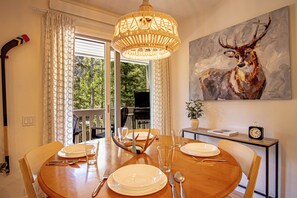 Un comedor con una mesa redonda, sillas, una araña y un cuadro de un ciervo, con vistas al exterior a través de la ventana.