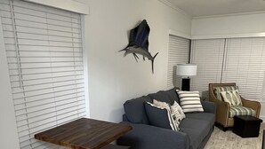 Een woonkamer met een houten tafel en bankje, een grijze bank met gestreepte kussens, een decoratieve vis aan de muur en een lamp op een bijzettafeltje.