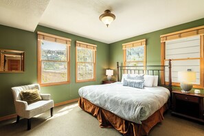 Une chambre avec un grand lit, une chaise, deux fenêtres, une table de chevet et une lampe.