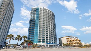 Un hotel alto y cilíndrico con fachada azul y blanca, rodeado de palmeras y una playa de arena.