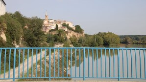 Vista del río de un pueblo con una iglesia destacada en una colina, un camino de tierra que lleva al pueblo, y una barandilla metálica azul en primer plano.