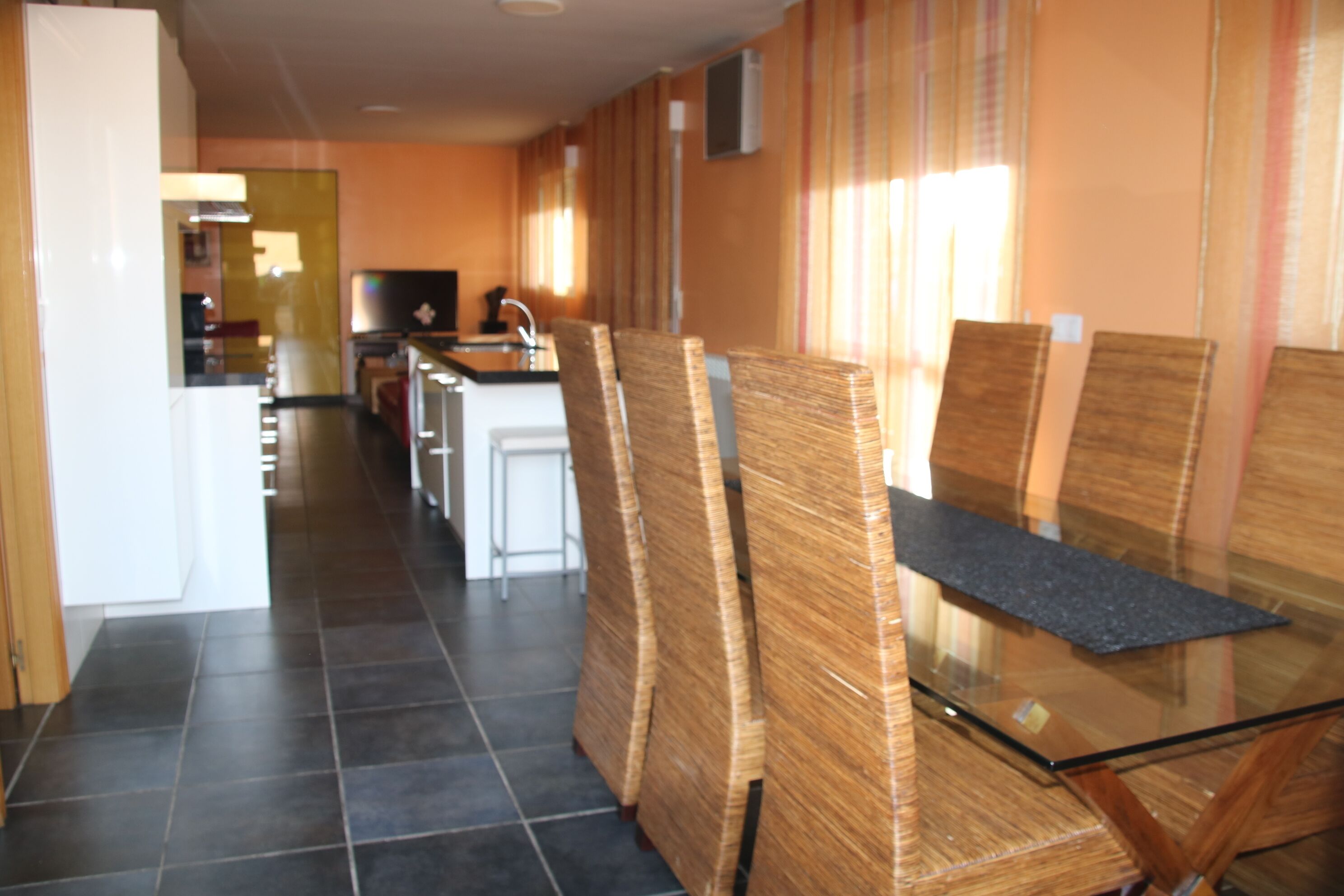 Un pasillo de hotel con una zona de cocina, una mesa de vidrio y sillas de mimbre.