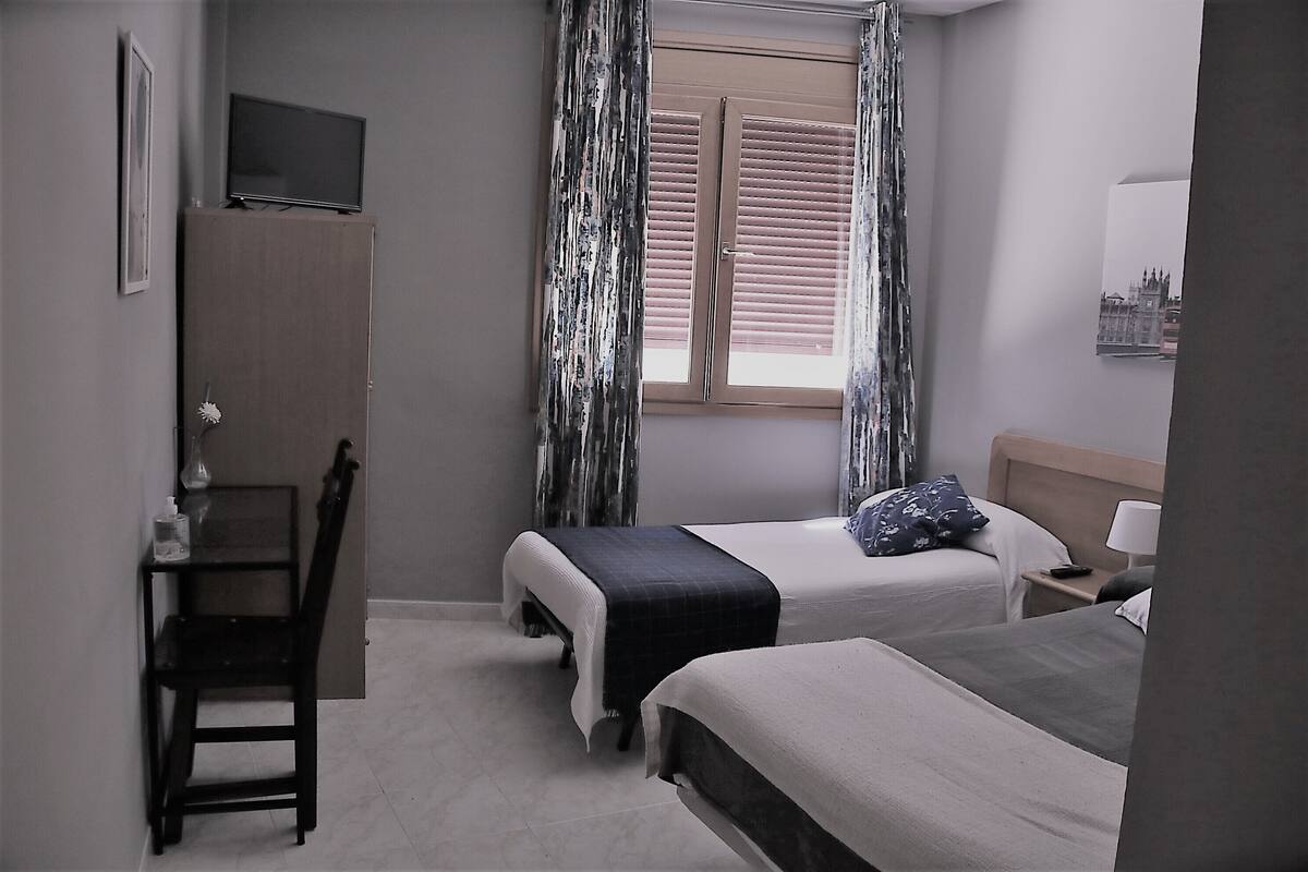 Ein Hotelzimmer mit zwei Betten, einem Schreibtisch, einem Stuhl, einem Fernseher und einem Fenster mit Vorhängen.