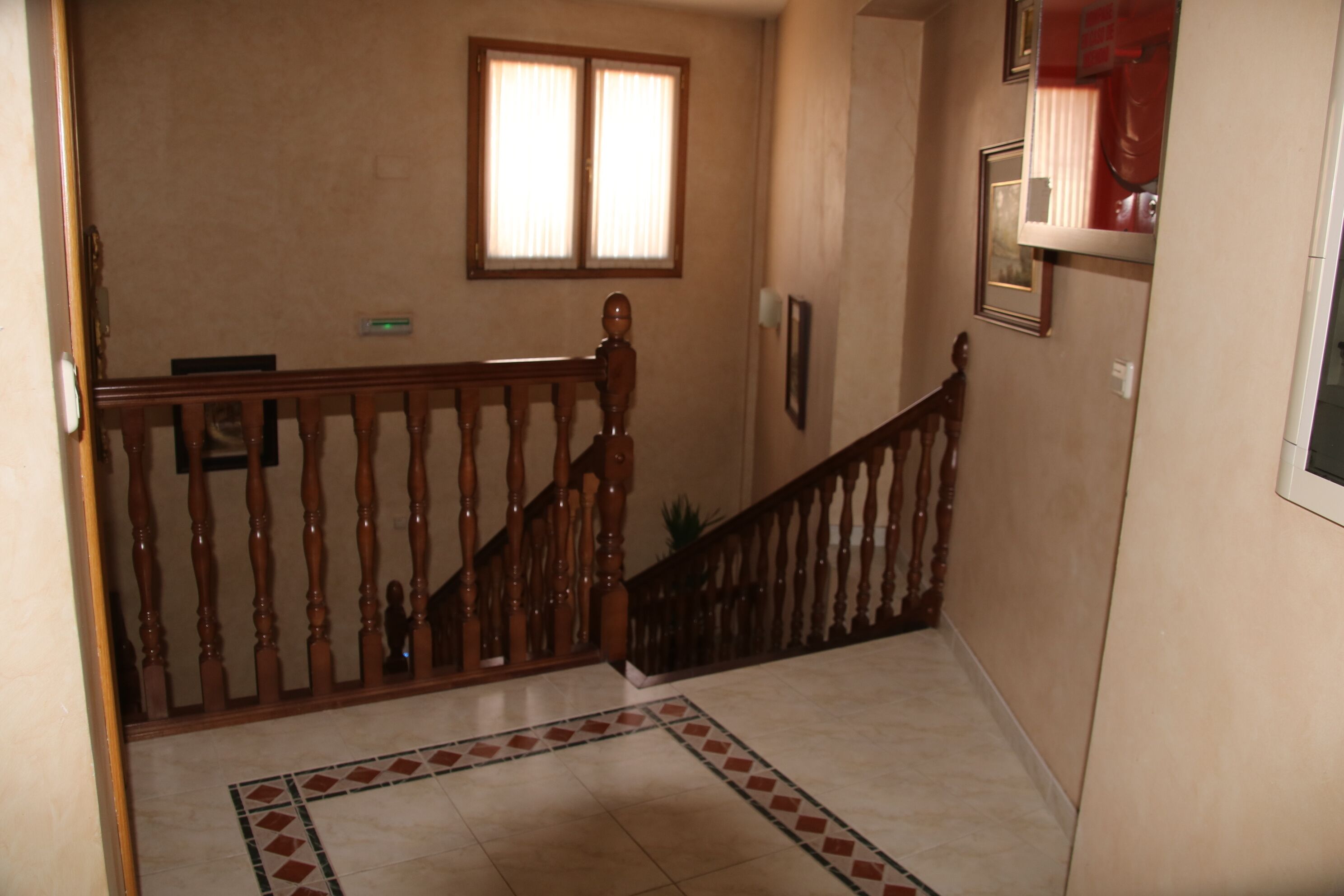 A hallway with a wooden staircase, a tiled floor, a window with blinds, and a door on the left.