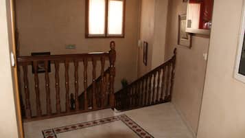 A hallway with a wooden staircase, a tiled floor, a window with blinds, and a door on the left.