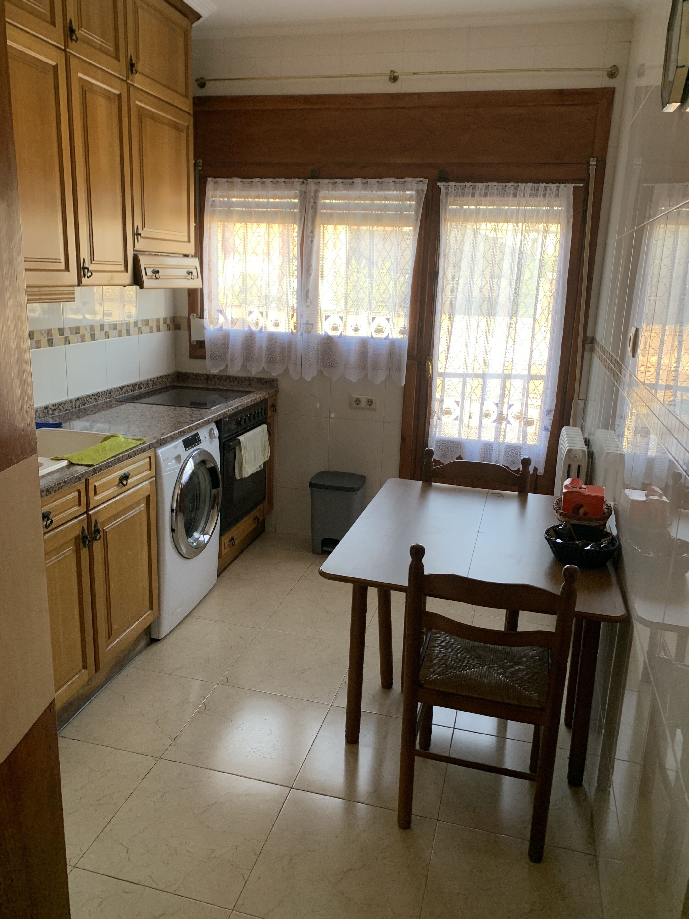 A compact kitchen with wooden cabinets, a built-in washing machine, a dining table with chairs, and a window with lace curtains.