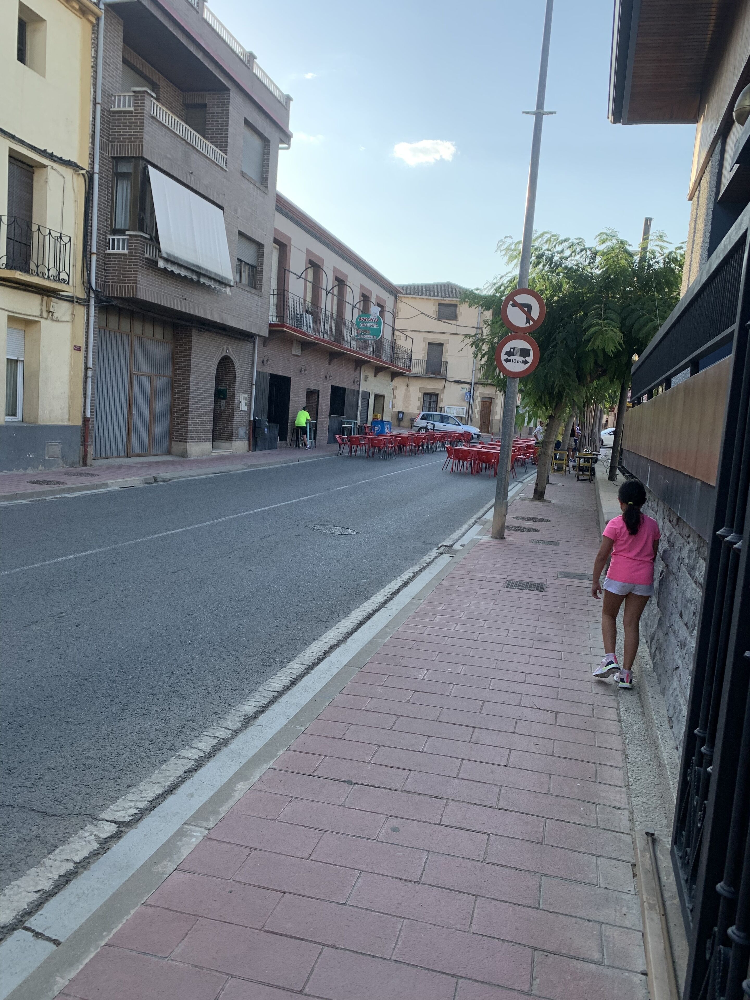 A street with a sidewalk, a pedestrian, and a row of outdoor tables and chairs.