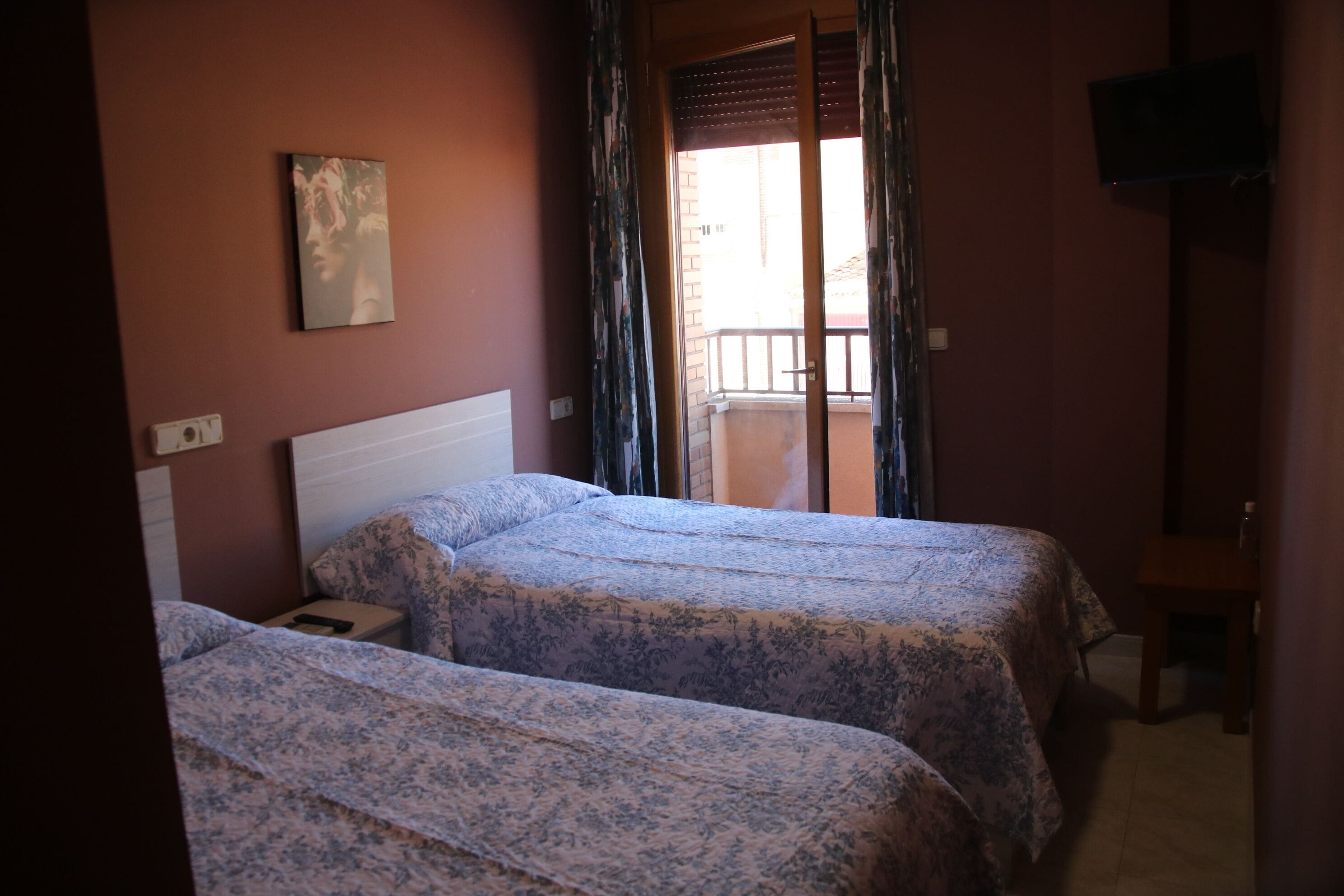 A room with two beds, a TV on the wall, and a balcony with curtains.