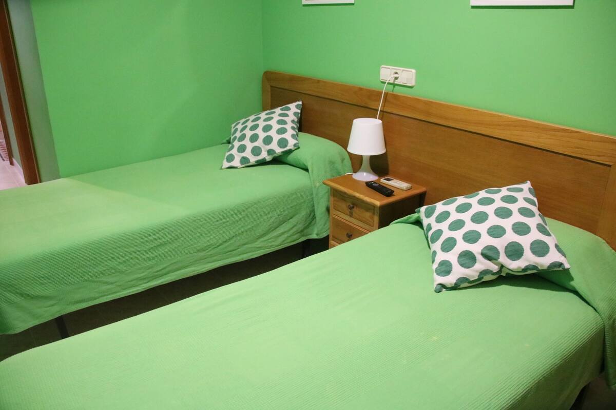 Ein Zimmer mit zwei Einzelbetten, jedes mit einer grünen Tagesdecke und einem gepunkteten Kissen. Zwischen den Betten steht ein Nachttisch aus Holz mit einer Lampe und einem Telefon.
