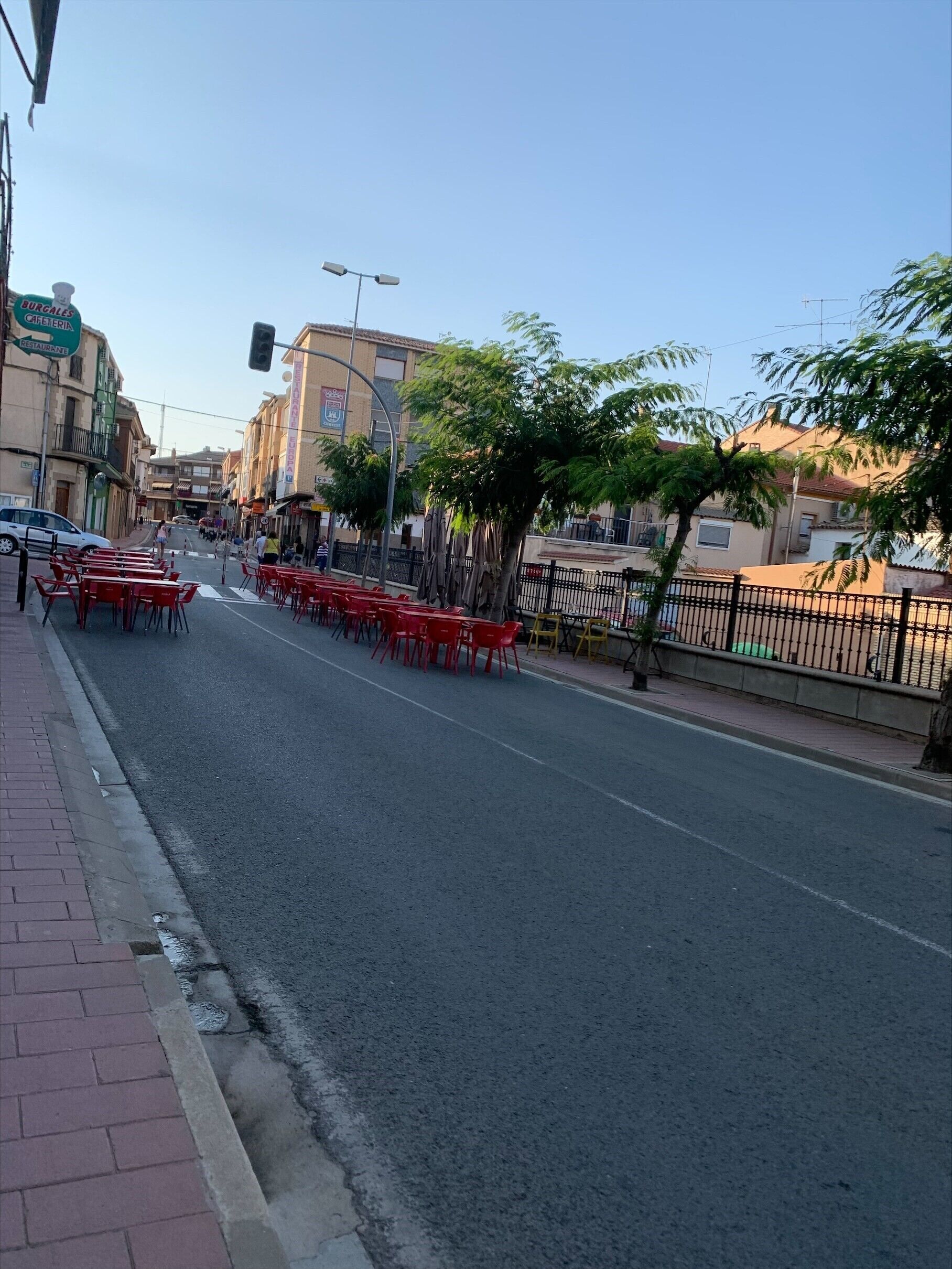 Una calle con sillas rojas alineadas a un lado, cielo despejado y edificios al fondo.