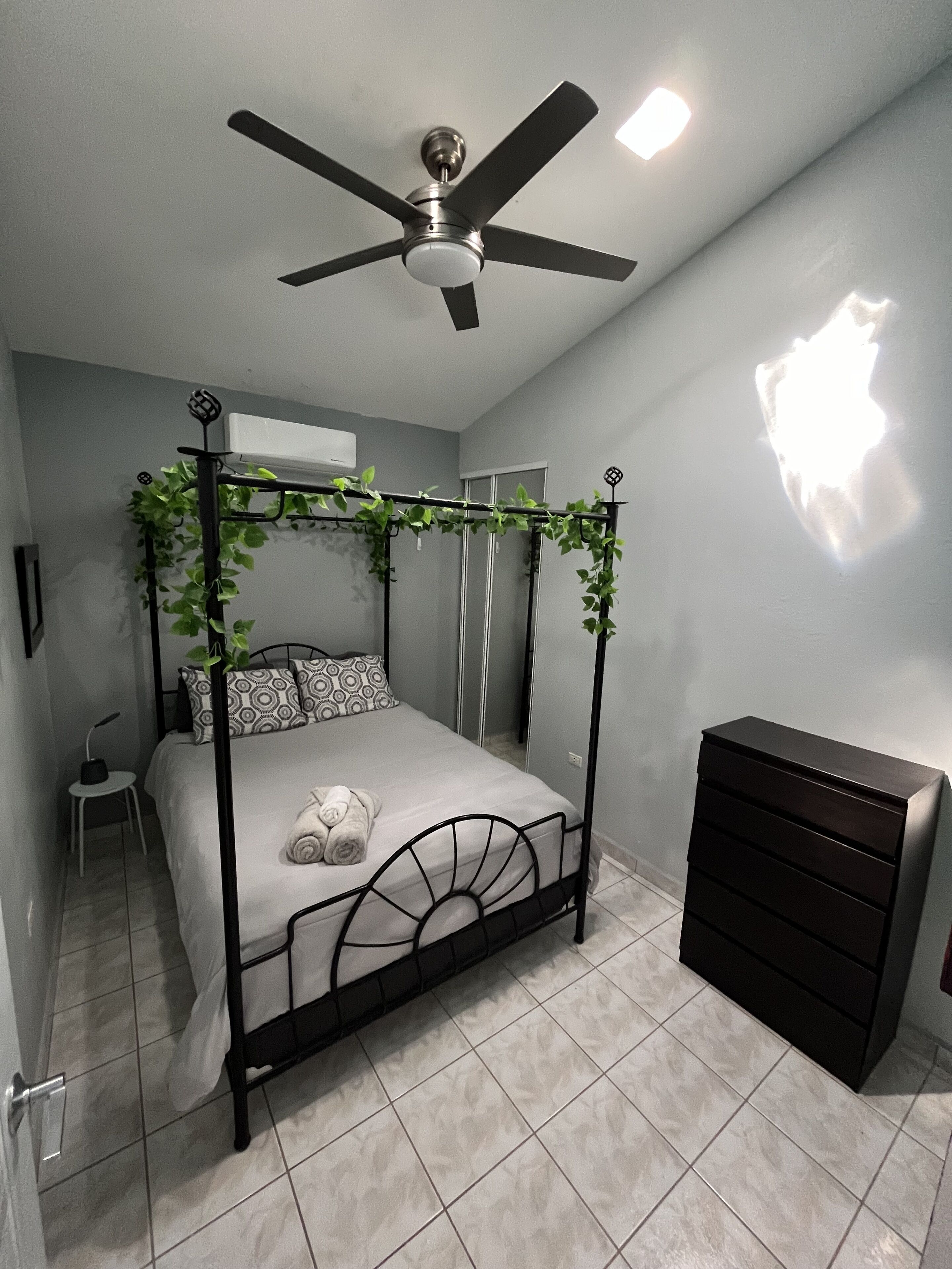 Une chambre à coucher avec un lit à baldaquin, un ventilateur de plafond, une commode et une petite table sur laquelle est posée une plante.