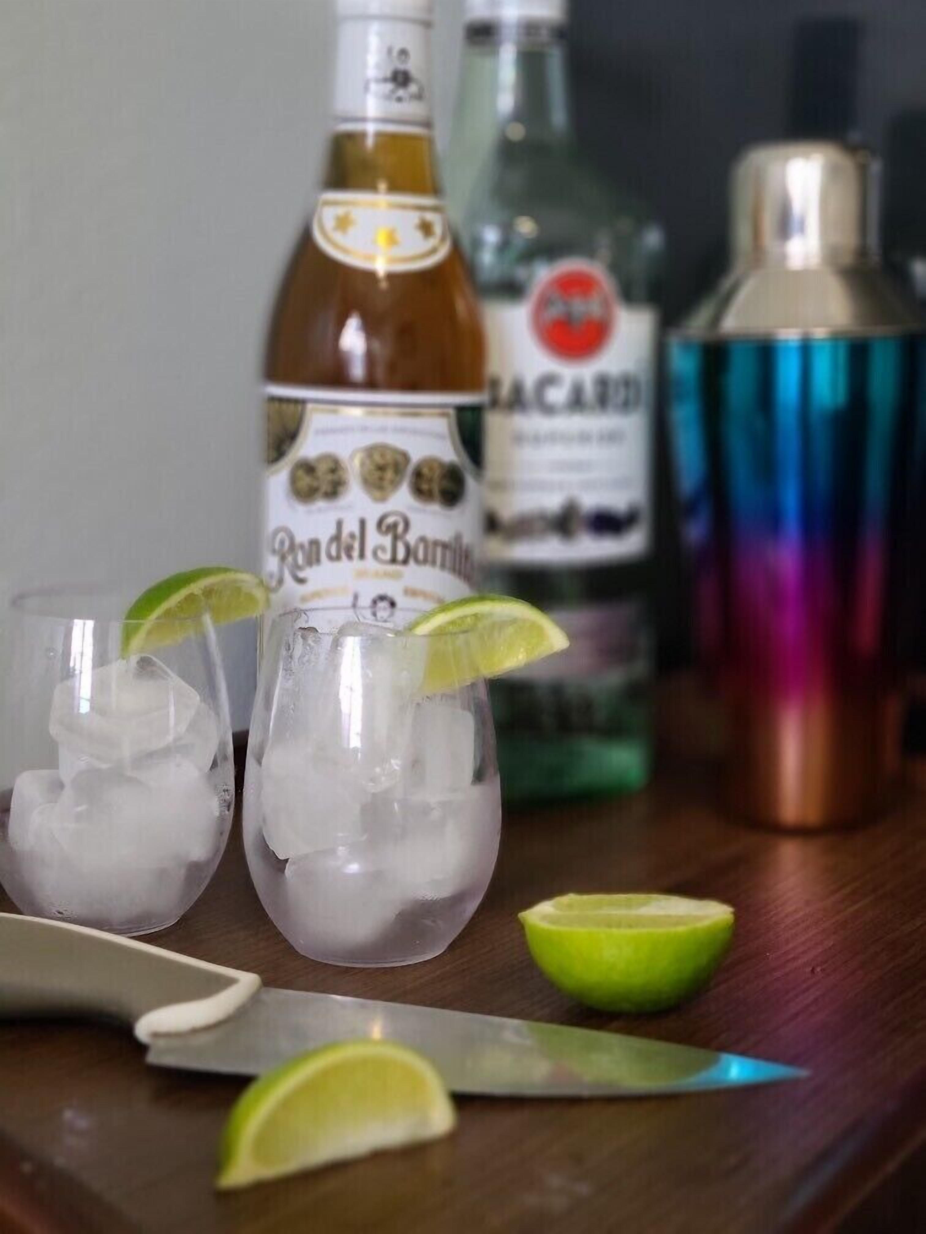 Deux verres remplis de glaçons et de quartiers de citron vert, une bouteille de « Randal Barrick », une bouteille de « Macaparo » et un verre à fond épais de couleur cuivrée, le tout posé sur une surface en bois.