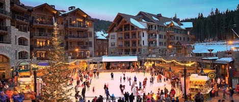 A bustling outdoor ice skating rink surrounded by multi-story buildings with balconies and large windows.