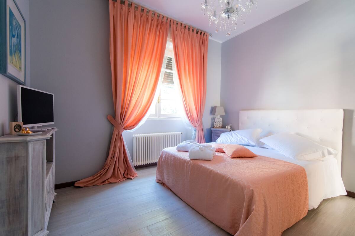 A bedroom with a bed, a nightstand, a lamp, a TV, and a window with curtains.