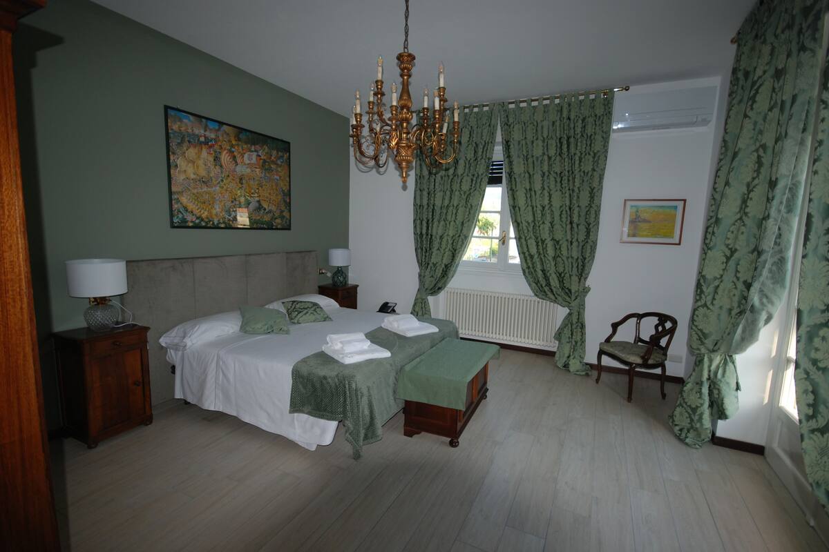 A bedroom with a large bed, a chandelier, green curtains, and a painting on the wall.