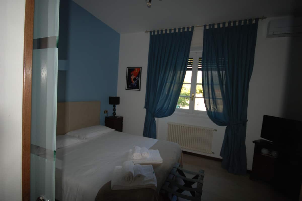 A bedroom with a bed, a nightstand, a window with curtains, a radiator, and a television.