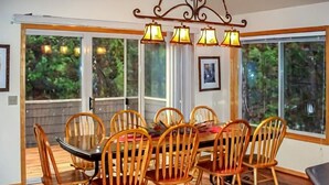 A dining area with a round table and six wooden chairs, large windows with curtains, and a chandelier. - NEW Mountain Home at the Deschutes River,Bikes,Game Room,Hot Tub,AC,SHARC Passes (Bend)