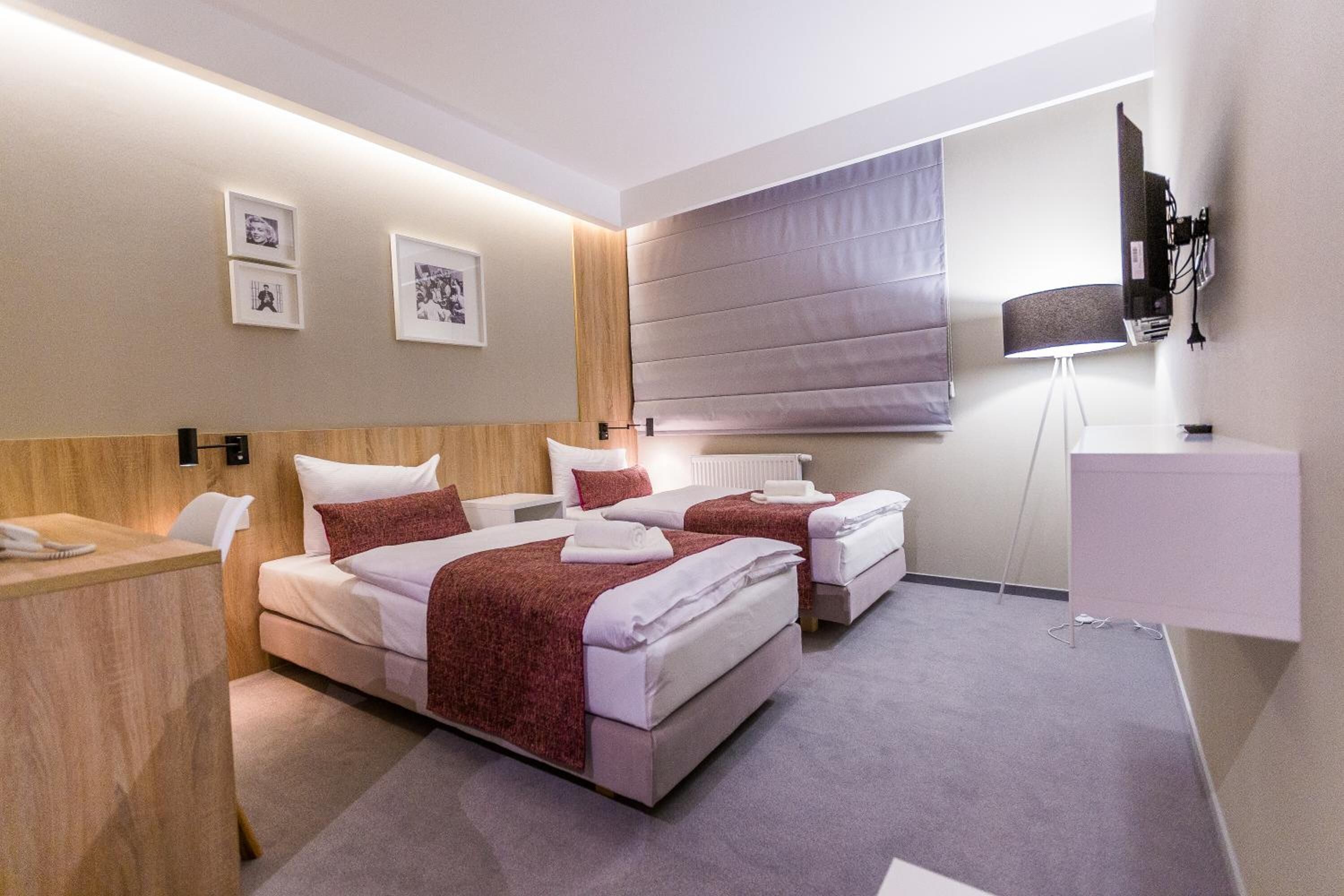 Una habitación de hotel moderna con dos camas, un escritorio, una lámpara de pie y un televisor montado en la pared.