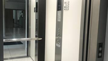 An elevator with metallic walls and a glass door, featuring a handrail and control panel with buttons.