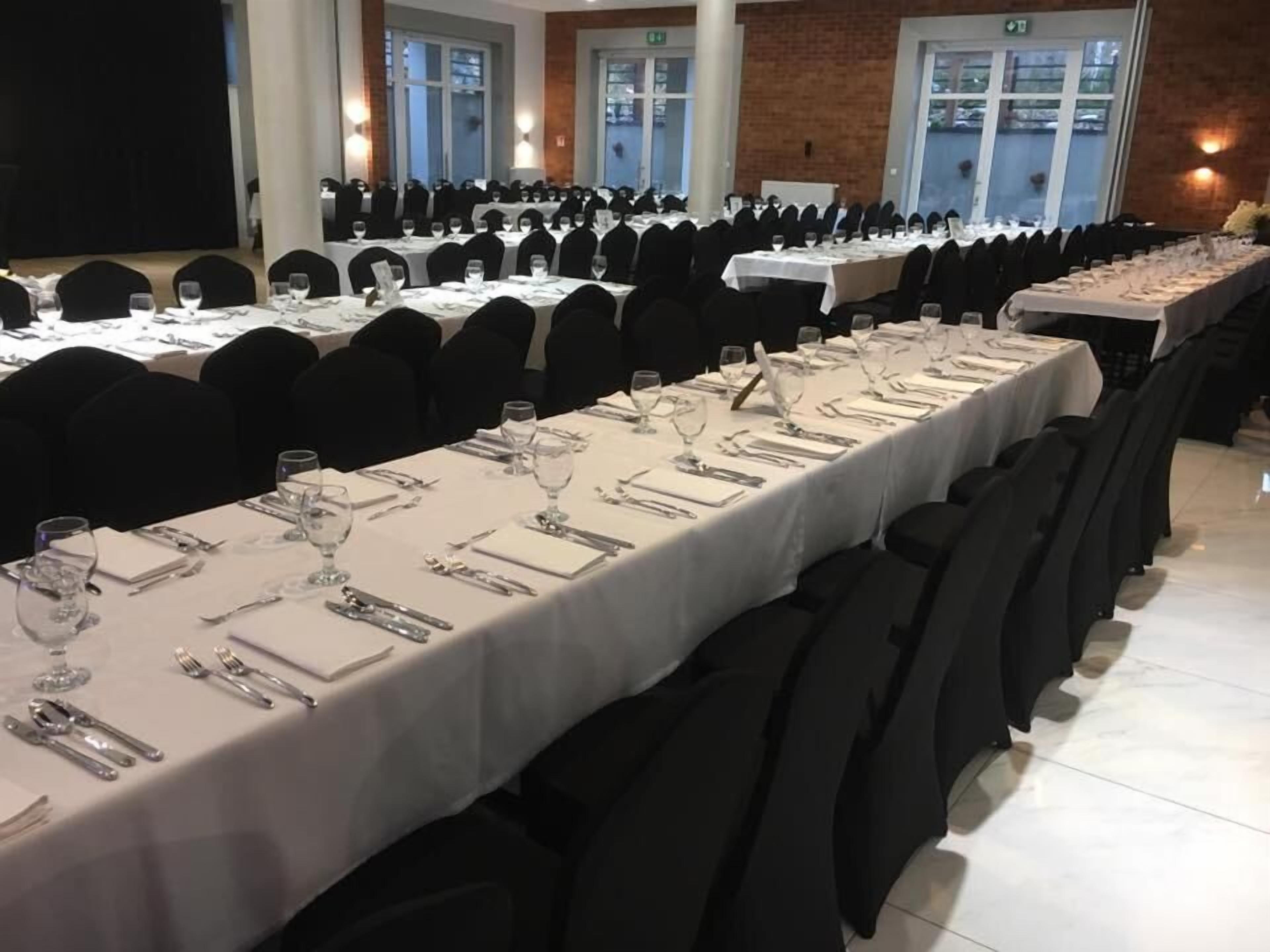 Een feestzaal met lange tafels gedekt voor een formeel evenement, met witte tafelkleden en zwarte stoelen.